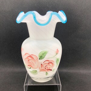 May include: White glass vase with a ruffled rim and a blue trim. The vase features hand-painted pink roses and green leaves. The vase has a bulbous body and a narrow neck.