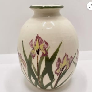 May include: A white ceramic vase with a green rim and a floral design. The vase is decorated with purple and yellow irises and green leaves.