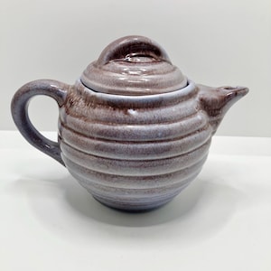 May include: A purple and brown ceramic teapot with a lid and a rounded body. The teapot has a textured, striped design.