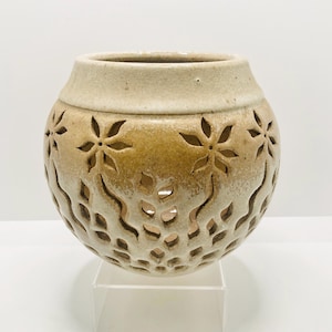 May include: A beige ceramic vase with a round shape and a wide opening. The vase features cut-out floral and wavy patterns, creating a decorative design. The top rim is a lighter shade of beige.