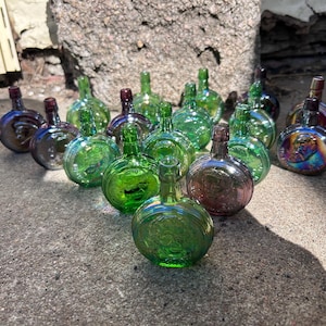 May include: A collection of small, round glass bottles in various colors, including green, purple, and iridescent hues. The bottles have a bulbous shape with a narrow neck and a small stopper. They are arranged on a rough, gray surface.