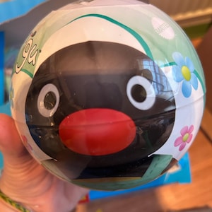 Puede incluir: Un juguete redondo con un personaje de dibujos animados de pingüino con una cara negra, ojos blancos y un pico rojo. El juguete está envuelto en plástico transparente y decorado con flores y la palabra "Yoga". El embalaje incluye la palabra "Mattel".