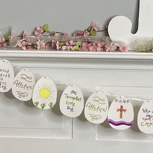 Könnte beinhalten: Ein weißes Papier-Osterei-Banner mit einer Reihe weißer Papiereier, die mit bunten Zeichnungen und Texten verziert sind. Die Eier haben Sätze wie "Jesus Christus ist heute auferstanden", "Halleluja", "Oh Trompete Heiliger Tag", "Wer hat einmal am Kreuz", und "Halleluja".