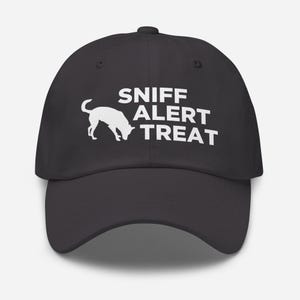 Op de afbeelding: Een zwarte baseball cap met de witte tekst "SNIFF ALERT TREAT" en een wit silhouet van een hond. De cap heeft een gebogen klep en een knoop aan de bovenkant. Ideaal voor hondenbezitters.