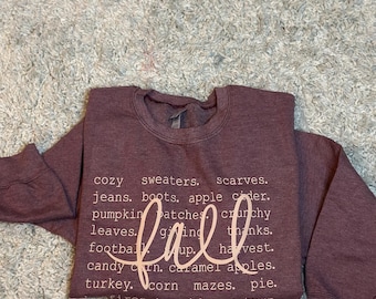 Puede incluir: Una sudadera granate con la palabra "fall" en escritura cursiva. La sudadera tiene una lista de palabras con temática otoñal, incluyendo "cozy sweaters", "pumpkin patches" y "caramel apples".