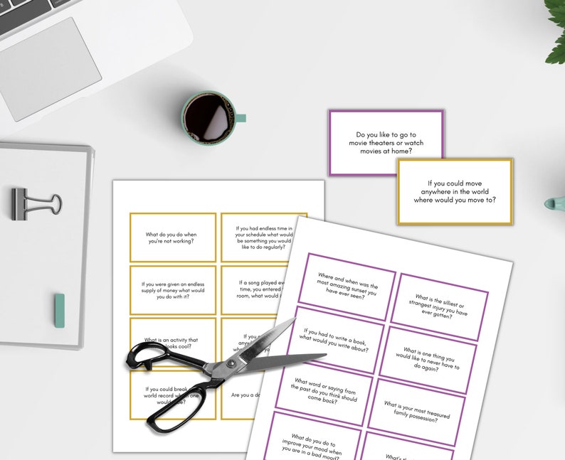 May include: A set of 12 gold and purple conversation starter cards with questions printed on them. The cards are on a white surface with a laptop, a cup of coffee, and a pair of scissors.