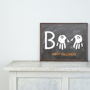 May include: A framed chalkboard art piece with the word "Boo" in white, featuring two handprint ghosts. The phrase "Happy Halloween" is written in orange below. The frame is dark brown, and the artwork is displayed on a white cabinet.