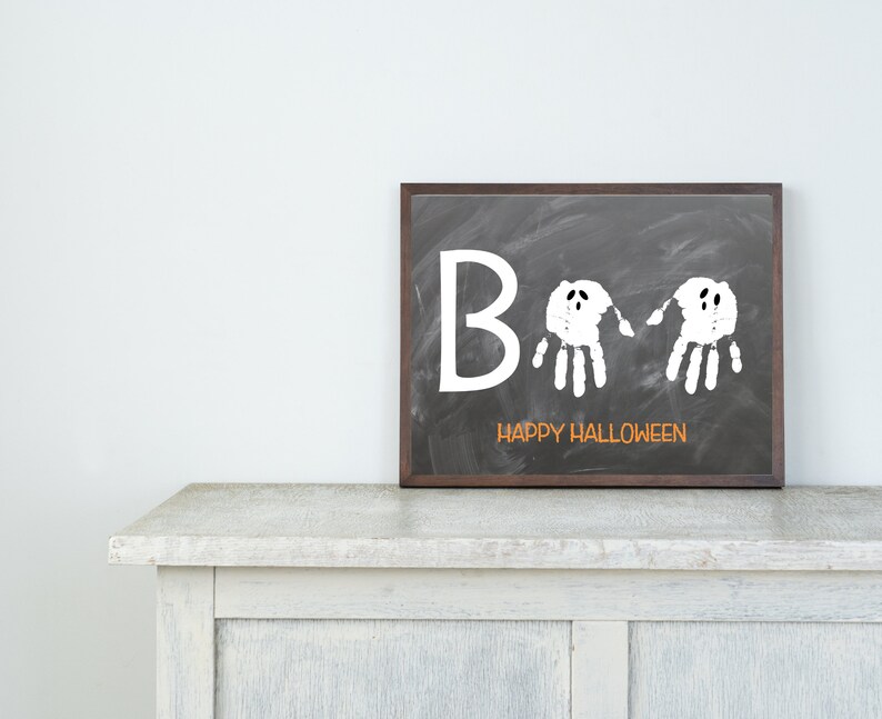 May include: A framed chalkboard art piece with the word "Boo" in white, featuring two handprint ghosts. The phrase "Happy Halloween" is written in orange below. The frame is dark brown, and the artwork is displayed on a white cabinet.