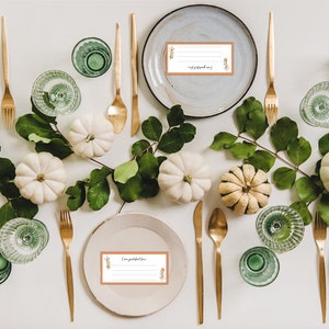 May include: A Thanksgiving table setting with white pumpkins, green leaves, gold silverware, and place cards that say "I am grateful for".