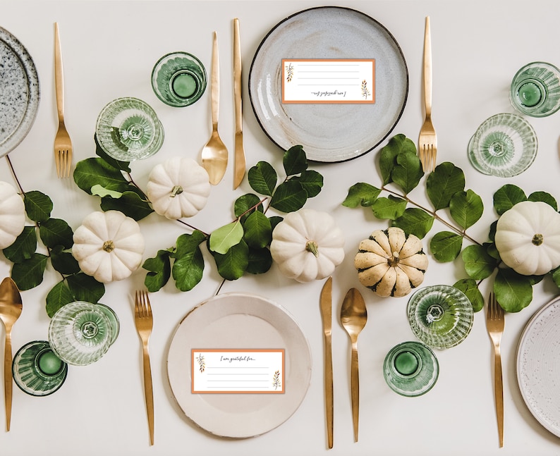 May include: A Thanksgiving table setting with white pumpkins, green leaves, gold silverware, and place cards that say "I am grateful for".