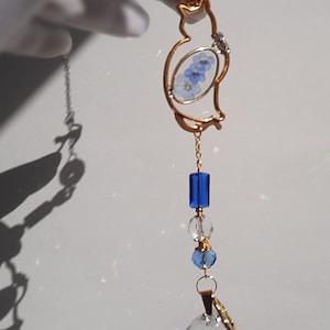 Può includere: Una silhouette di gatto in metallo dorato con fiori blu incapsulati in resina appesa a una catena con perline di cristallo blu e trasparenti e un grande prisma di cristallo trasparente.