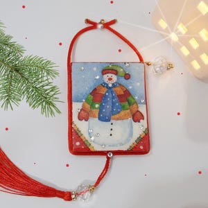 Op de afbeelding: Een rode en witte kerstversiering met een sneeuwpop met een blauwe sjaal en rode wanten. De versiering is versierd met glitter en heeft een rode kwast.