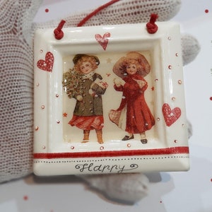 Op de afbeelding: Witte keramische ornament met een rode lijst en rode harten. De ornament heeft een vintage afbeelding van twee meisjes in rode en groene jurken die een kerstboom en een kleine witte vogel vasthouden. De ornament heeft het woord "Happy" onderaan geschreven.