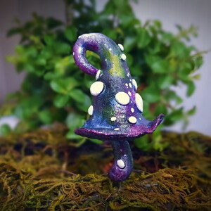 Puede incluir: Un hongo caprichoso de color morado y verde con lunares blancos, perfecto para un jardín de hadas o una escena en miniatura.