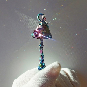 Può includere: Un ciondolo a forma di fungo fantasioso con una finitura iridescente viola e verde. Il ciondolo presenta una grande gemma trasparente al centro del cappello del fungo.