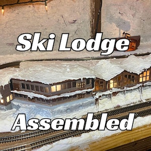 May include: A model train set featuring a ski lodge with snow-covered roofs and windows. The lodge is assembled and ready to be placed on the track. The text "Ski Lodge Assembled" is visible in the image.