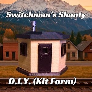 May include: A model kit of a small, beige, hexagonal building with a dark brown door and windows. The building is labeled "Switchman's Shanty D.I.Y. (Kit Form)" and is set against a backdrop of mountains and trees.