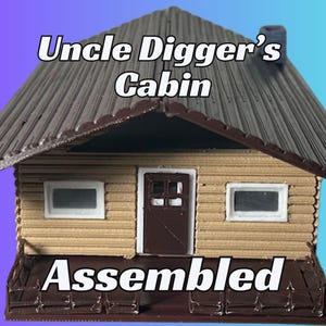May include: A small, assembled wooden cabin model with a brown roof, brown door, and two white windows. The cabin has a brown wooden porch with a railing. The text "Uncle Digger's Cabin" is above the cabin and the text "Assembled" is below the cabin.