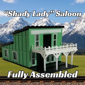 May include: A fully assembled green wooden model of a saloon building with a white balcony and the text "Shady Lady" Saloon. The model is set against a backdrop of snow-capped mountains and a grassy field.