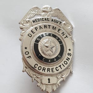 Puede incluir: Una insignia de metal plateado con un águila en la parte superior. El texto dice MEDICAL ASS'T DEPARTMENT OF CORRECTIONS STATE OF TEXAS. Un emblema circular central muestra una estrella rodeada por una corona, con el número 1 abajo.