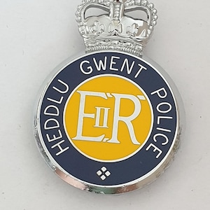 Puede incluir: Una insignia circular de la policía de Gwent con una corona plateada en la parte superior. El centro es amarillo con las letras ER y el número romano II. Un borde azul oscuro rodea el centro con el texto HEDDLU GWENT POLICE y un pequeño rombo.