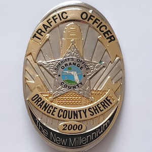 Könnte beinhalten: Ein ovales Metallabzeichen in Gold- und Silbertönen. Der Text lautet TRAFFIC OFFICER, ORANGE COUNTY SHERIFF und The New Millennium. Ein zentrales Sternemblem zeigt den Text SHERIFF'S OFFICE ORANGE COUNTY sowie das Jahr 2000.