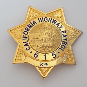 Puede incluir: Una insignia de estrella de seis puntas en tono dorado con el texto CALIFORNIA HIGHWAY PATROL en letras azules. El centro muestra un relieve del sello del estado de California con los números 6 1 5 y el texto K9 en una cinta curva inferior.