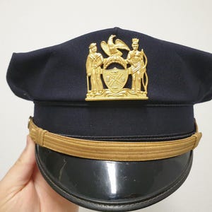 Può includere: Un berretto di servizio blu scuro con un vistoso emblema in metallo dorato sul davanti. L'emblema raffigura due figure che fiancheggiano uno scudo centrale sormontato da un'aquila. Un sottogola dorato poggia sopra una visiera nera lucida.