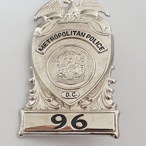 Peut inclure: Un insigne argenté de la police métropolitaine du district de Columbia avec un aigle au sommet, le texte 'METROPOLITAN POLICE', 'DISTRICT OF COLUMBIA', 'JUSTITIA OMNIBUS', 'D.C.' et le numéro '96' gravé en noir sur la partie inférieure.