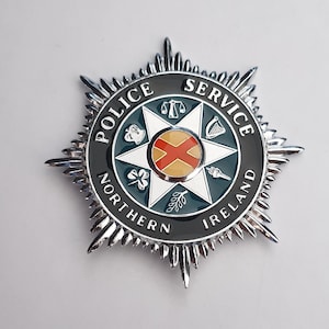 Könnte beinhalten: Ein silberfarbenes Metallabzeichen mit sternförmigem Design und einem dunkelgrünen kreisförmigen Rand. Der Text POLICE SERVICE NORTHERN IRELAND ist in Weiß eingraviert. In der Mitte befindet sich ein weißer Stern mit Symbolen wie Waage, Harfe, Fackel und Kleeblatt, die ein rotes Kreuz auf gelbem Grund umgeben.