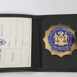 Puede incluir: Una cartera de cuero negro que contiene una tarjeta de identificación y una insignia de metal dorado. La tarjeta presenta el texto N.Y.P.D. y espacios para datos de identificación. La insignia muestra el texto City of New York Police Lieutenant.