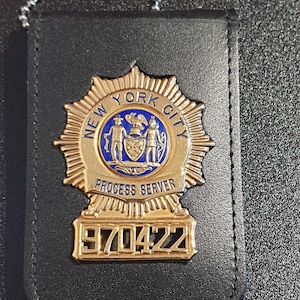 Könnte beinhalten: Ein goldfarbenes Metallabzeichen mit einer blauen kreisförmigen Mitte, die das Siegel von New York City, den Text "NEW YORK CITY" und "PROCESS SERVER" sowie die Nummer "970422" an der Basis zeigt. Das Abzeichen ist auf einem schwarzen Lederhalter befestigt.