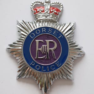 May include: A silver-toned Dorset Police badge featuring a central purple circle with the letters ER in silver. A blue circular border contains the text DORSET POLICE. The badge is topped with a silver crown featuring red enamel detailing.