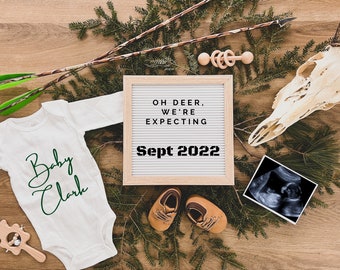 May include: A white onesie with the text "Baby Clark" in green script. A letter board with the text "Oh Deer, We're Expecting Sept 2022" is on a wooden surface with a deer skull, a wooden rattle, and a baby ultrasound image.