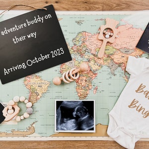Może przedstawiać: Tablica z napisem "New adventure buddy on their way Arriving October 2023" leży na mapie świata. Są dwa paszporty, body dla dziecka z napisem "Baby Bing" i zdjęcie USG.