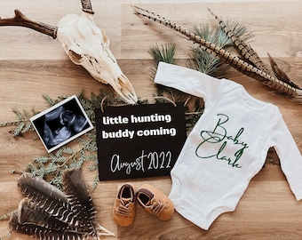 May include: A black chalkboard sign with the text "little hunting buddy coming August 2022". A white onesie with the text "Baby Clark" in green script. A small ultrasound image, a deer skull, and feathers are arranged around the sign and onesie.