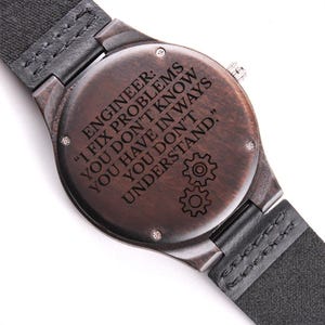 May include: A wooden watch with a dark brown face and a black leather strap. The watch face has the engraved text: "ENGINEER: I FIX PROBLEMS YOU DON'T KNOW YOU HAVE IN WAYS YOU DON'T UNDERSTAND."