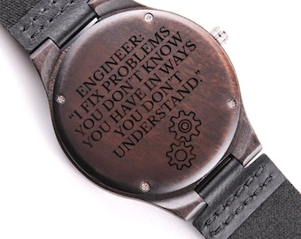 May include: A wooden watch with a dark brown face and a black leather strap. The watch face has the engraved text: "ENGINEER: I FIX PROBLEMS YOU DON'T KNOW YOU HAVE IN WAYS YOU DON'T UNDERSTAND."