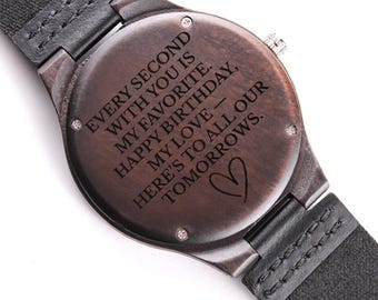 May include: A wooden wristwatch with a dark brown face and a black leather strap. The watch face features engraved text: "EVERY SECOND WITH YOU IS MY FAVORITE. HAPPY BIRTHDAY, MY LOVE - HERE'S TO ALL OUR TOMORROWS." A heart symbol is also engraved.