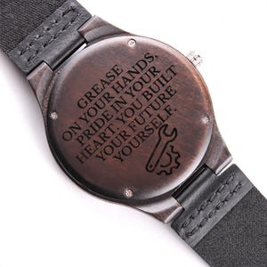 May include: A wooden watch with a dark brown face and a black leather strap. The watch face has the engraved text: "GREASE ON YOUR HANDS, PRIDE IN YOUR HEART. YOU BUILT YOUR FUTURE YOURSELF."