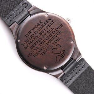 Peut inclure: Une montre en bois avec un cadran marron foncé et un bracelet en cuir noir. Le cadran de la montre comporte le texte gravé : "LE TRAVAIL QUE VOUS FAITES EST INVISIBLE POUR BEAUCOUP, MAIS RESSENTI PROFONDÉMENT PAR TOUS. MERCI."