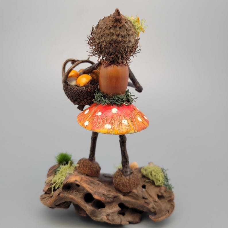 May include: A whimsical fairy figurine crafted from natural materials. The fairy wears a red mushroom skirt with white spots and carries a basket of orange fruits. The figure is posed on a piece of driftwood, with moss accents.
