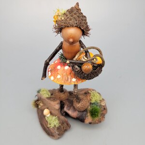 May include: A whimsical figurine of a woodland sprite. The figure has a gourd body, twig arms, and a mushroom skirt with white polka dots. It carries a basket filled with miniature mushrooms and sits on a wooden base with moss.