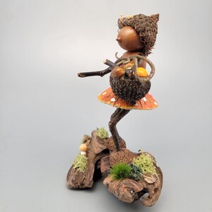 May include: A whimsical figurine of a woodland sprite carrying a basket of mushrooms. The figure has a brown acorn cap hat and a mushroom-shaped skirt. The base is made of wood and moss, creating a naturalistic scene.
