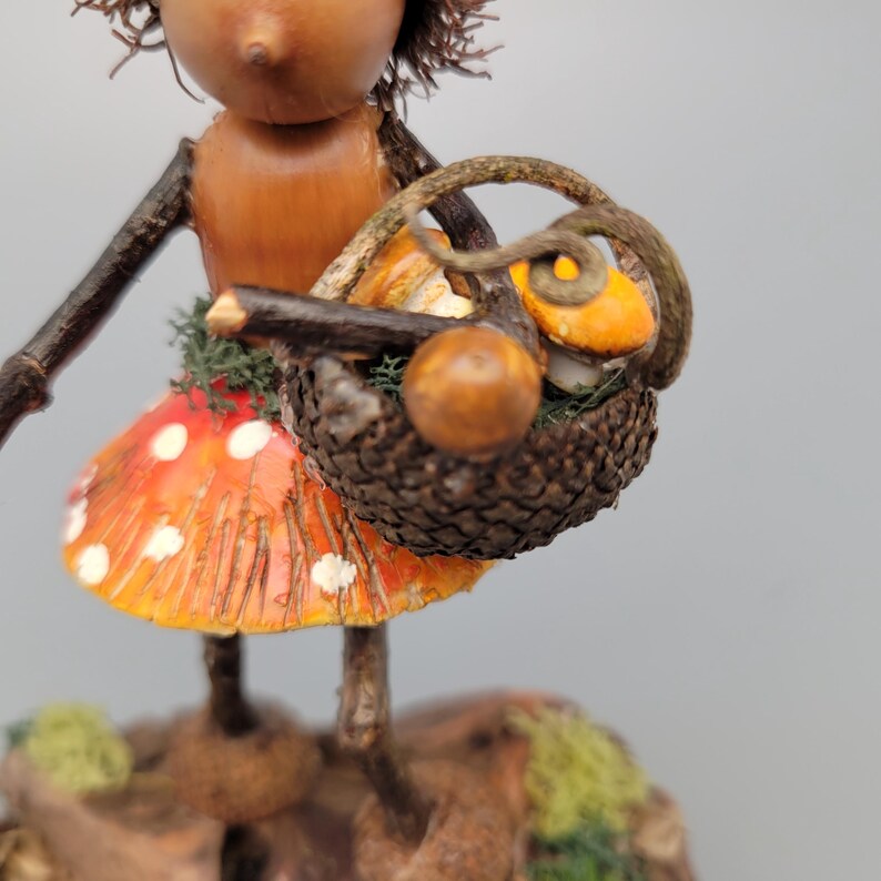 May include: A whimsical figurine of a fairy with a mushroom skirt and a basket. The fairy's skirt is orange with white spots. The basket is filled with miniature pumpkins and mushrooms. The fairy is made of wood and acorn caps.