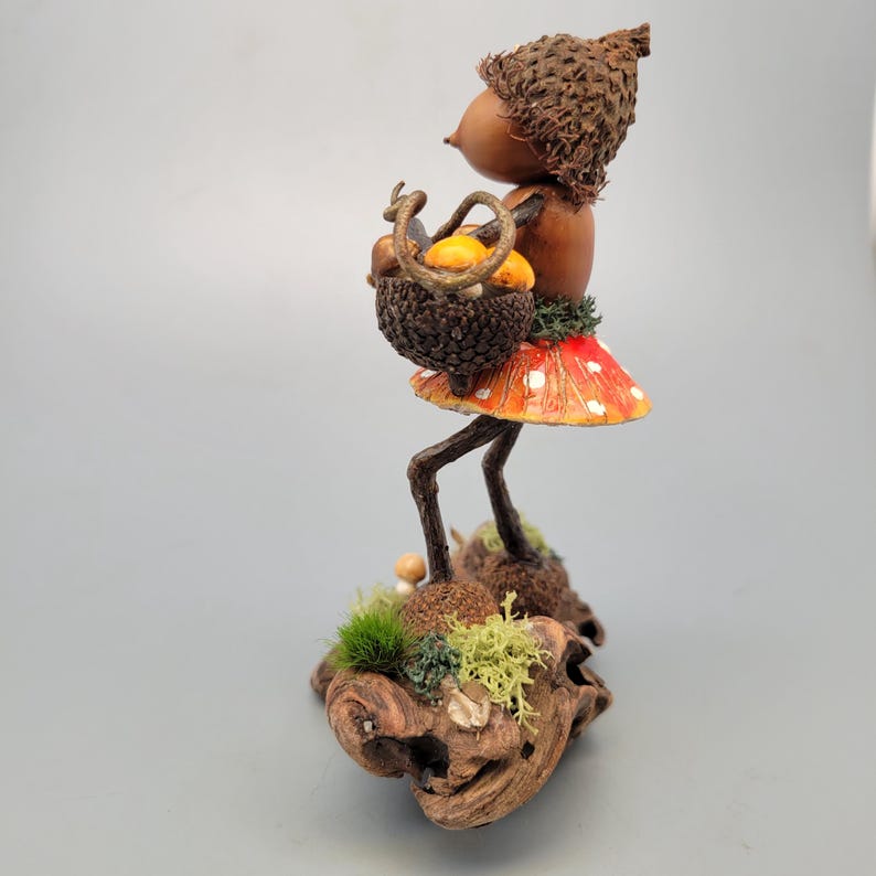 May include: A whimsical acorn figurine with a brown cap and body, holding a basket of mushrooms. It wears a red and white spotted mushroom skirt and stands on a wooden base with moss and lichen. The figurine is a unique decorative item.
