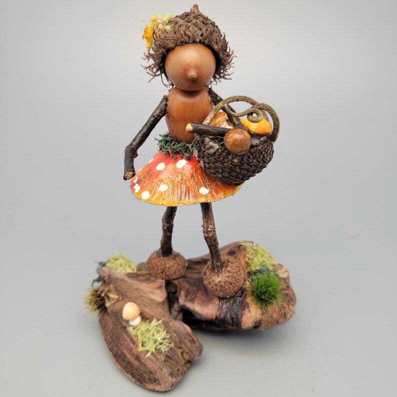 May include: A whimsical wooden figurine of a fairy-like creature. It has a brown acorn cap head, twig arms and legs, and a mushroom skirt. The fairy holds a basket with miniature mushrooms and stands on a wooden base with moss.
