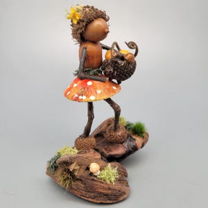 May include: A whimsical figurine of a woodland fairy. The fairy has a brown acorn head, twig arms and legs, and a red mushroom skirt with white spots. She carries a basket of fruit and stands on a piece of wood with moss.