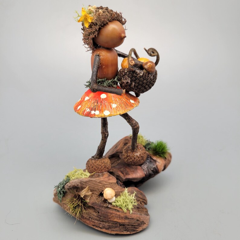 May include: A whimsical figurine of a woodland fairy. The fairy has a brown acorn head, twig arms and legs, and a red mushroom skirt with white spots. She carries a basket of fruit and stands on a piece of wood with moss.
