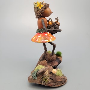 May include: A whimsical figurine of a woodland creature with a mushroom skirt and a basket. The figure is made of natural materials, with a red and white spotted mushroom skirt and a head adorned with a flower. The figurine is standing on a piece of wood.